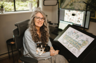 Christina Merkley holding her puppy Presley and sitting in front of her drawing tablet with a group of course attendes on the nearby screen.